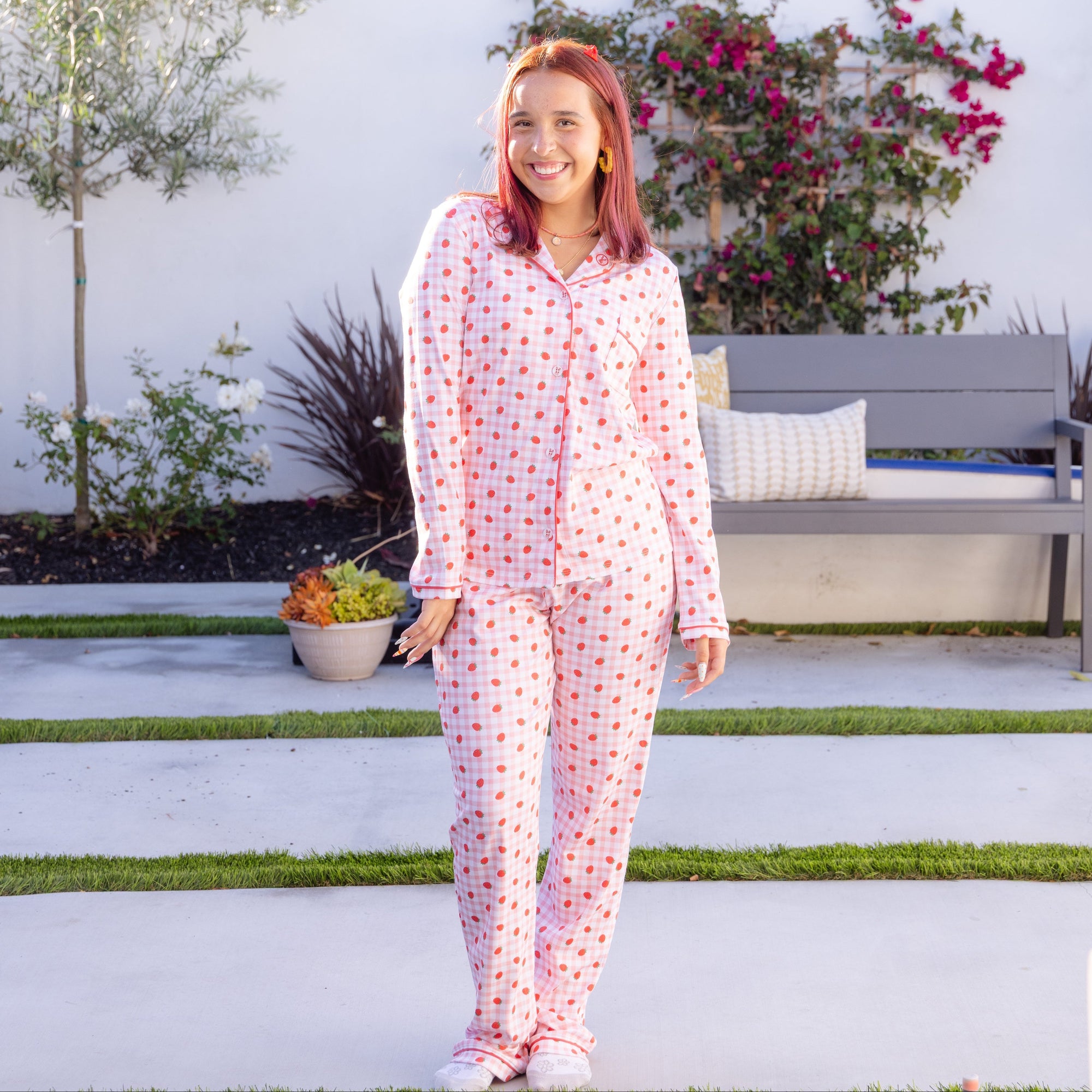 A woman stands outdoors wearing a matching long-sleeve pajama set with a strawberry pattern and a pink gingham pattern