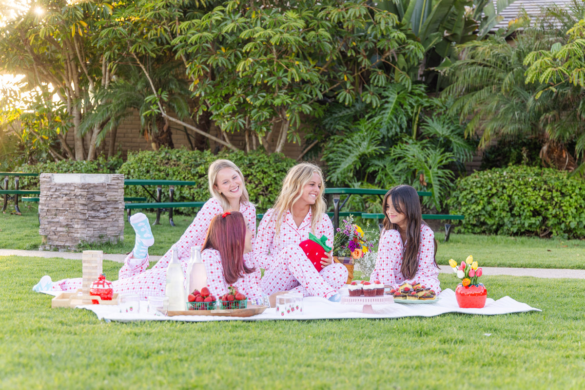 Three girls in matching pink gingham and strawberry print pajamas sit on a white blanket having a picnic outdoors on a green lawn, surrounded by food, flowers, and lush trees.
