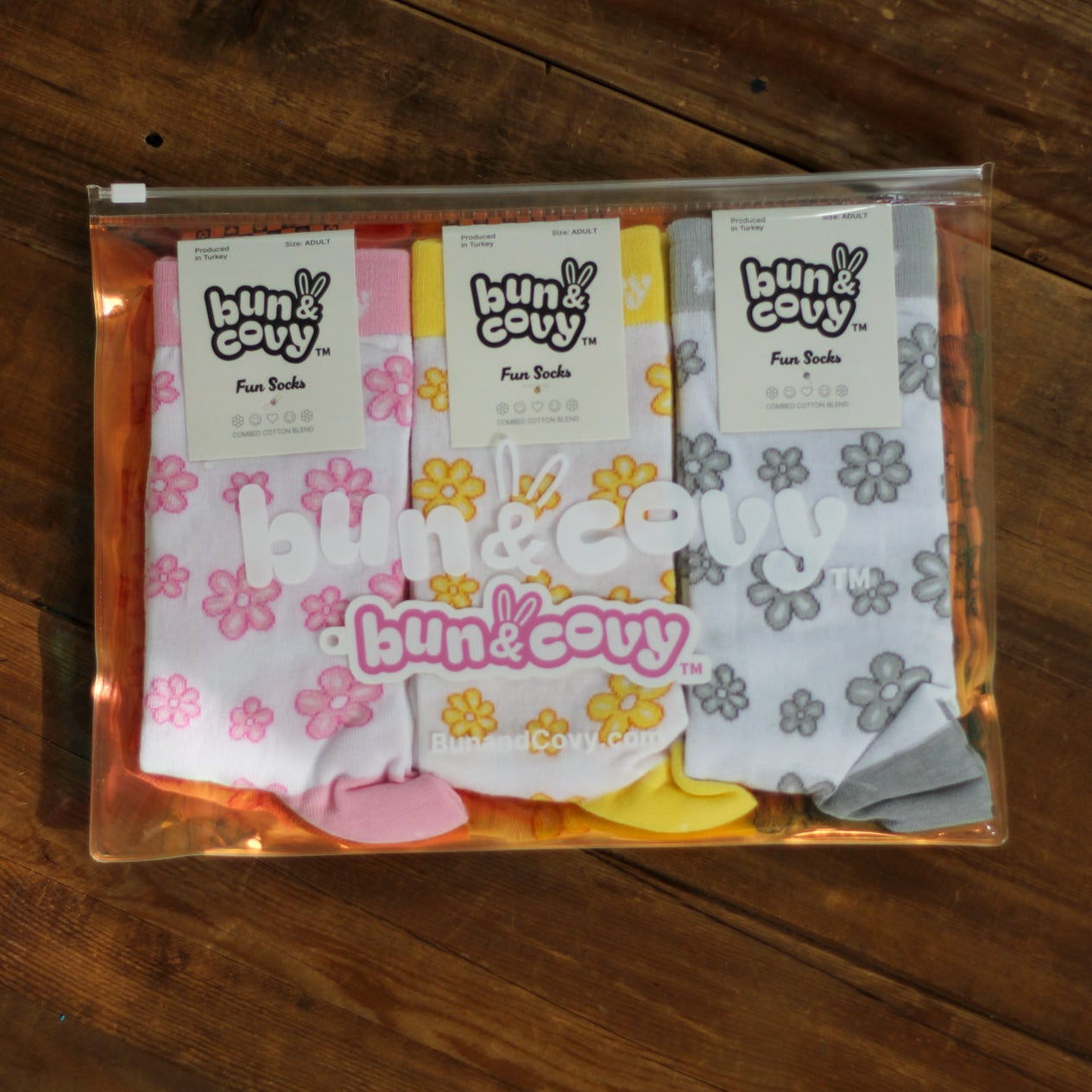 A clear zippered pouch containing three pairs of Bun & Covy branded knit socks, displayed on a dark wood plank surface. The socks are white with colorful floral patterns in pink, yellow, and gray, and each pair has its own branded paper header card.