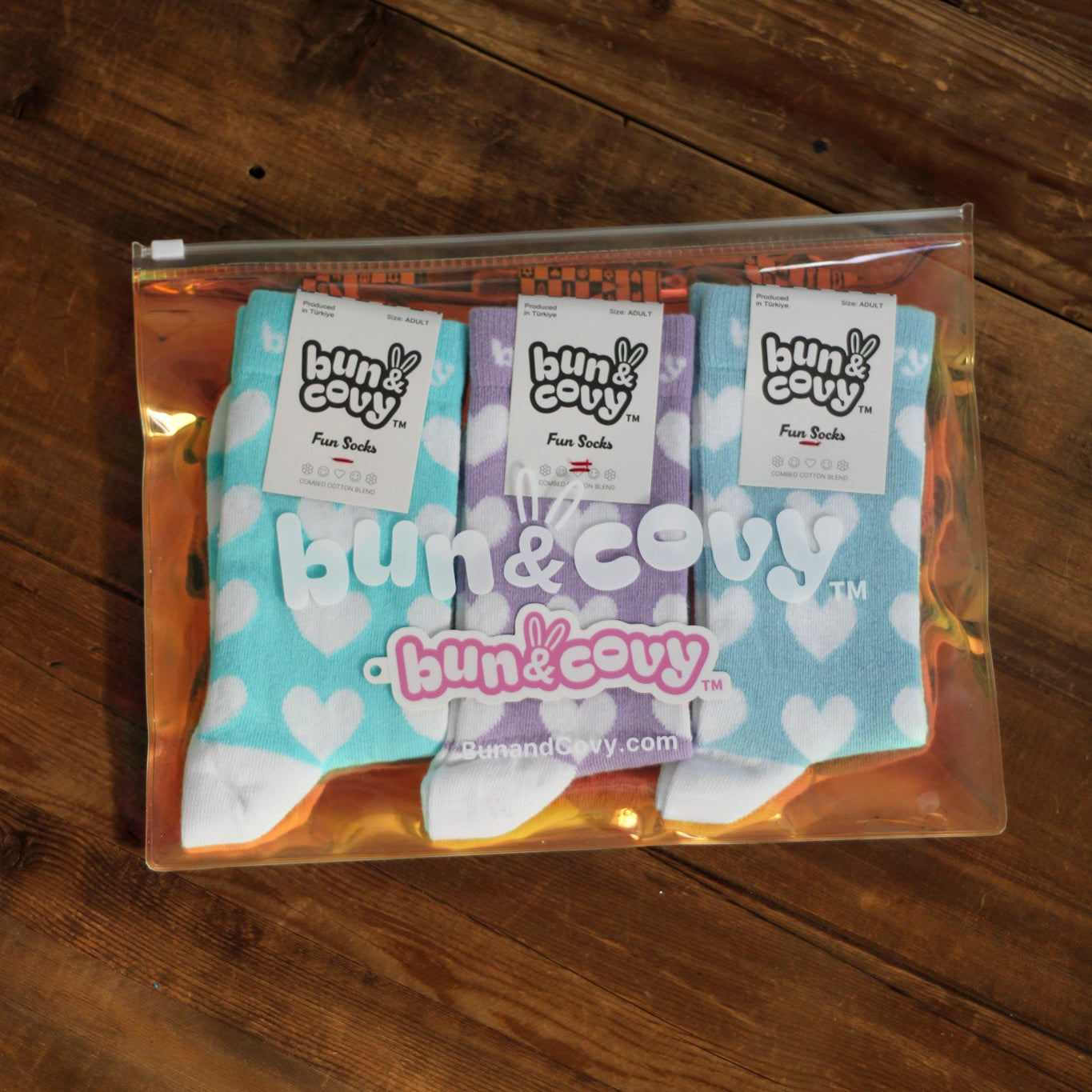 Three pairs of Bun & Covy socks in a clear zippered pouch, displayed on a dark wooden surface. The socks feature light blue, lavender purple, and white colors with heart patterns, and each has a white paper label with the Bun & Covy logo.