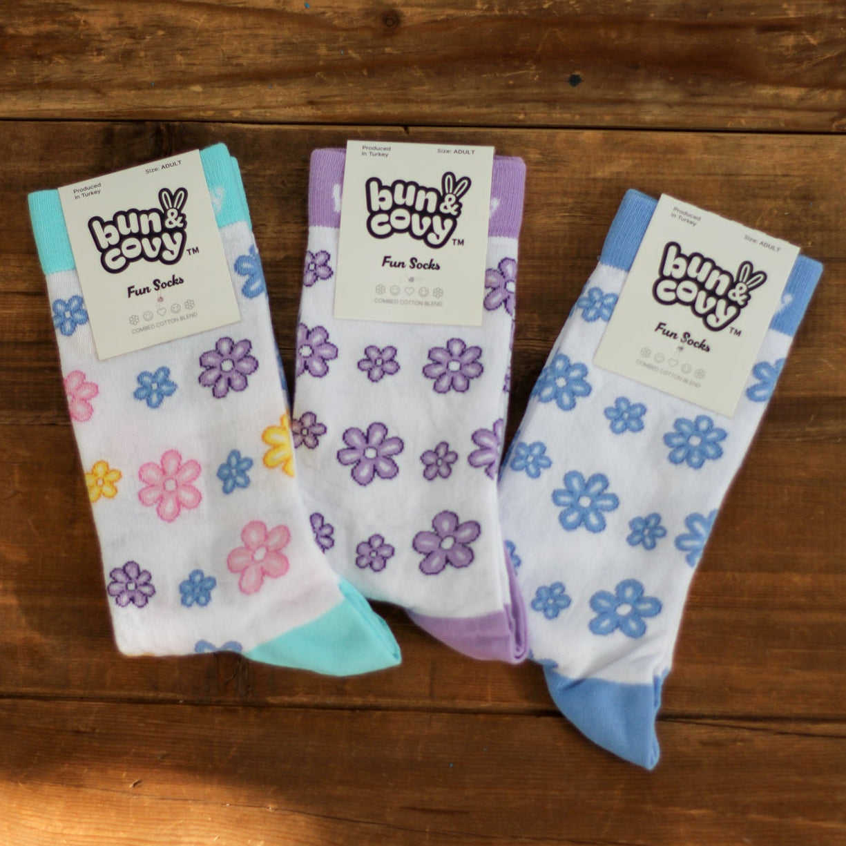 Three pairs of white crew socks with colorful floral patterns, displayed on a wooden surface. Each pair has a paper tag attached that reads 'bun & covy Fun Socks'.