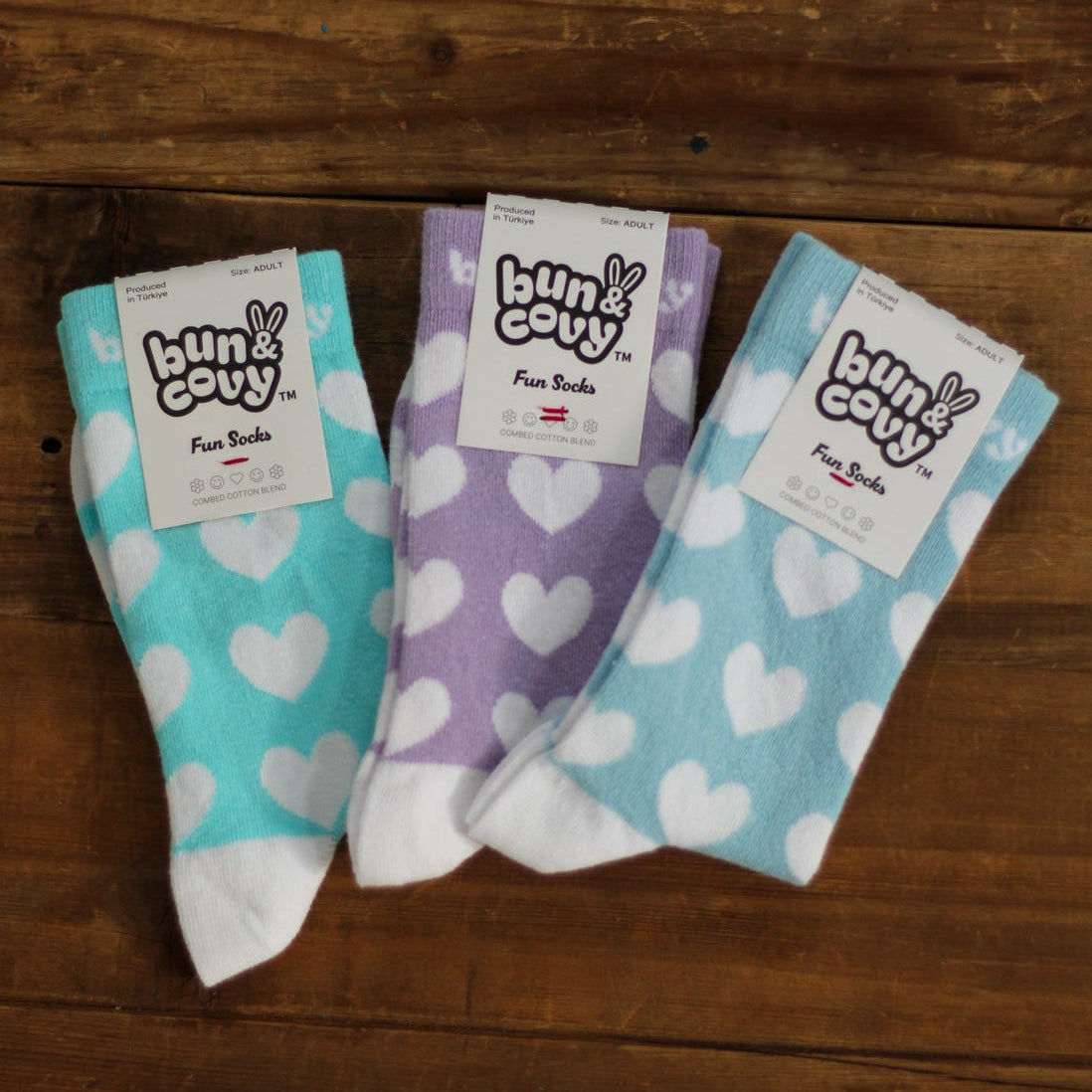 Three pairs of heart-patterned socks from Bun & Covy are displayed on a rustic wooden background. The left pair is teal with white hearts, the middle pair is lavender with white hearts, and the right pair is light blue with white hearts.