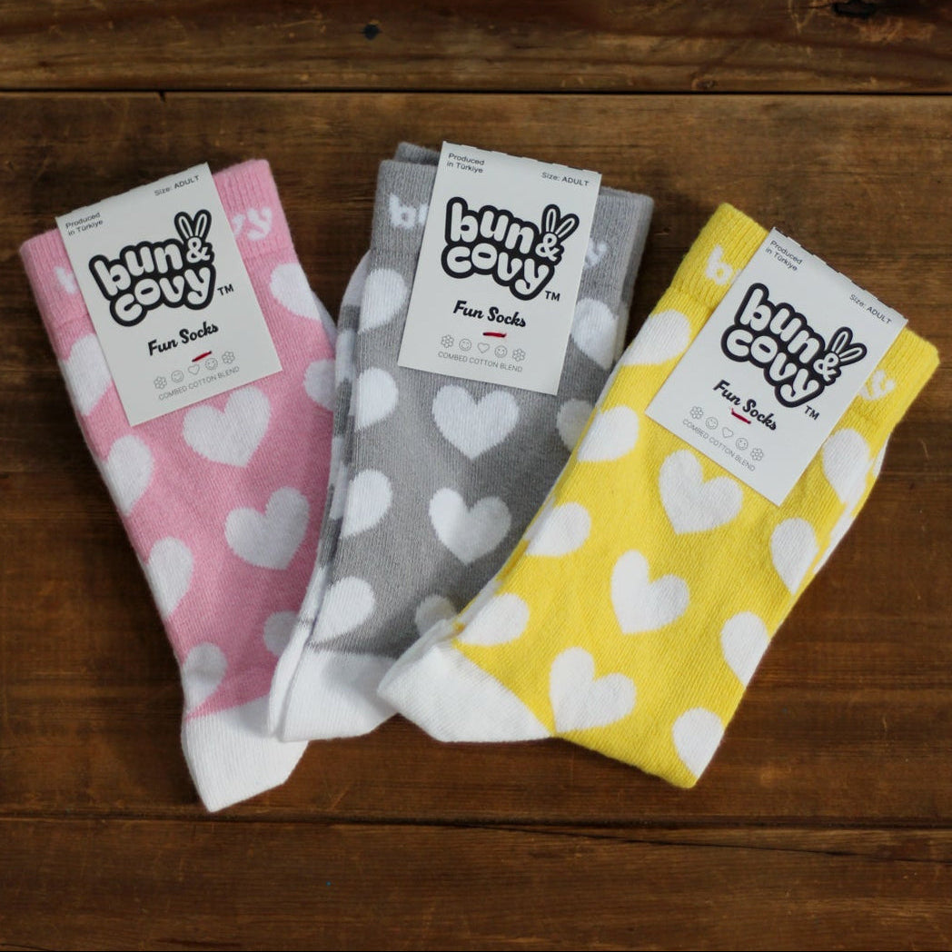 Three pairs of patterned crew socks laid flat on a dark wood surface: pink with white hearts, gray with white hearts, and yellow with white hearts. Each pair has a white paper tag with the brand name 'Bun & Covy' visible.