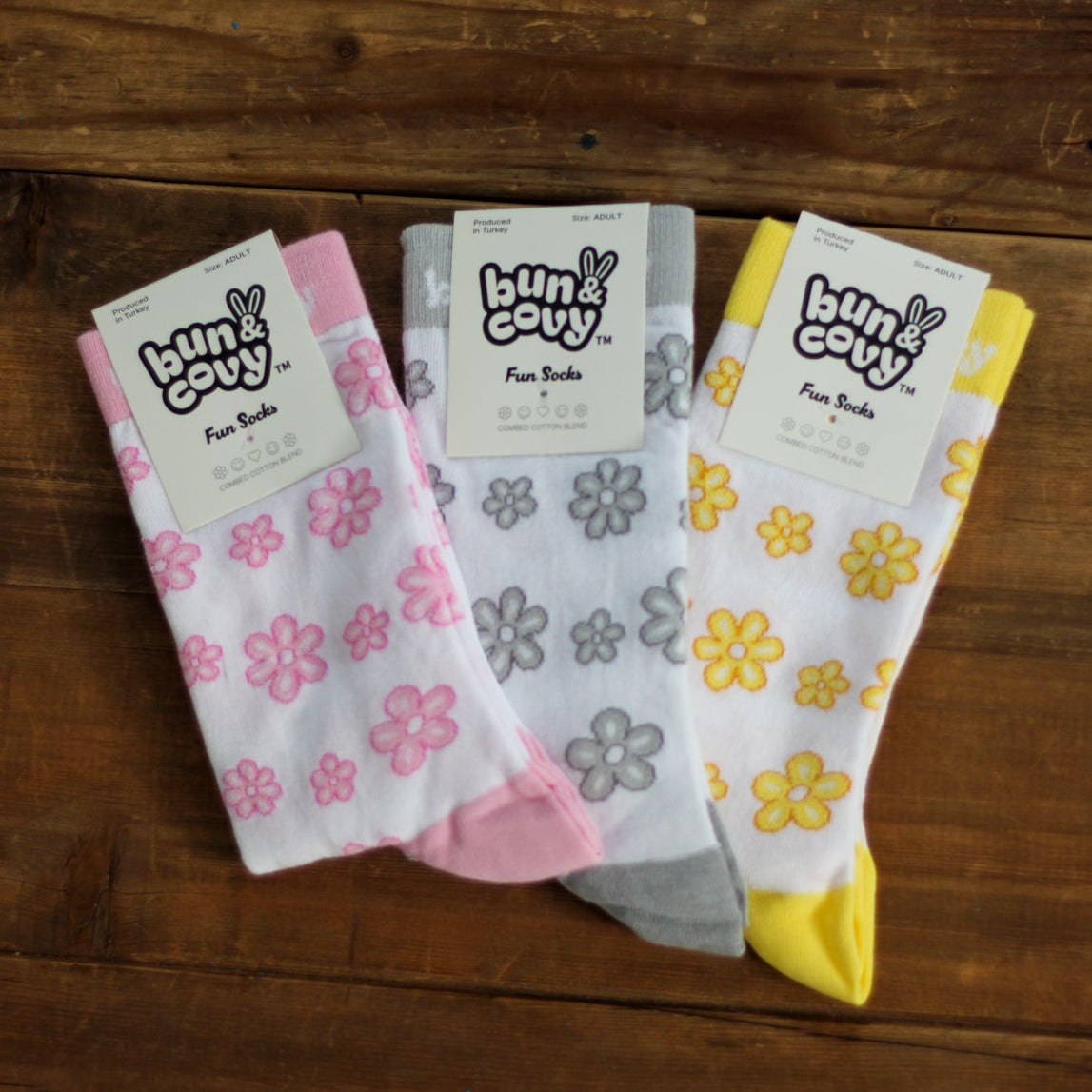 Three pairs of floral crew socks in pink, gray, and yellow, displayed on a dark wooden surface. Each pair has a white paper header card with the brand 'Bun & Covy' printed on it.
